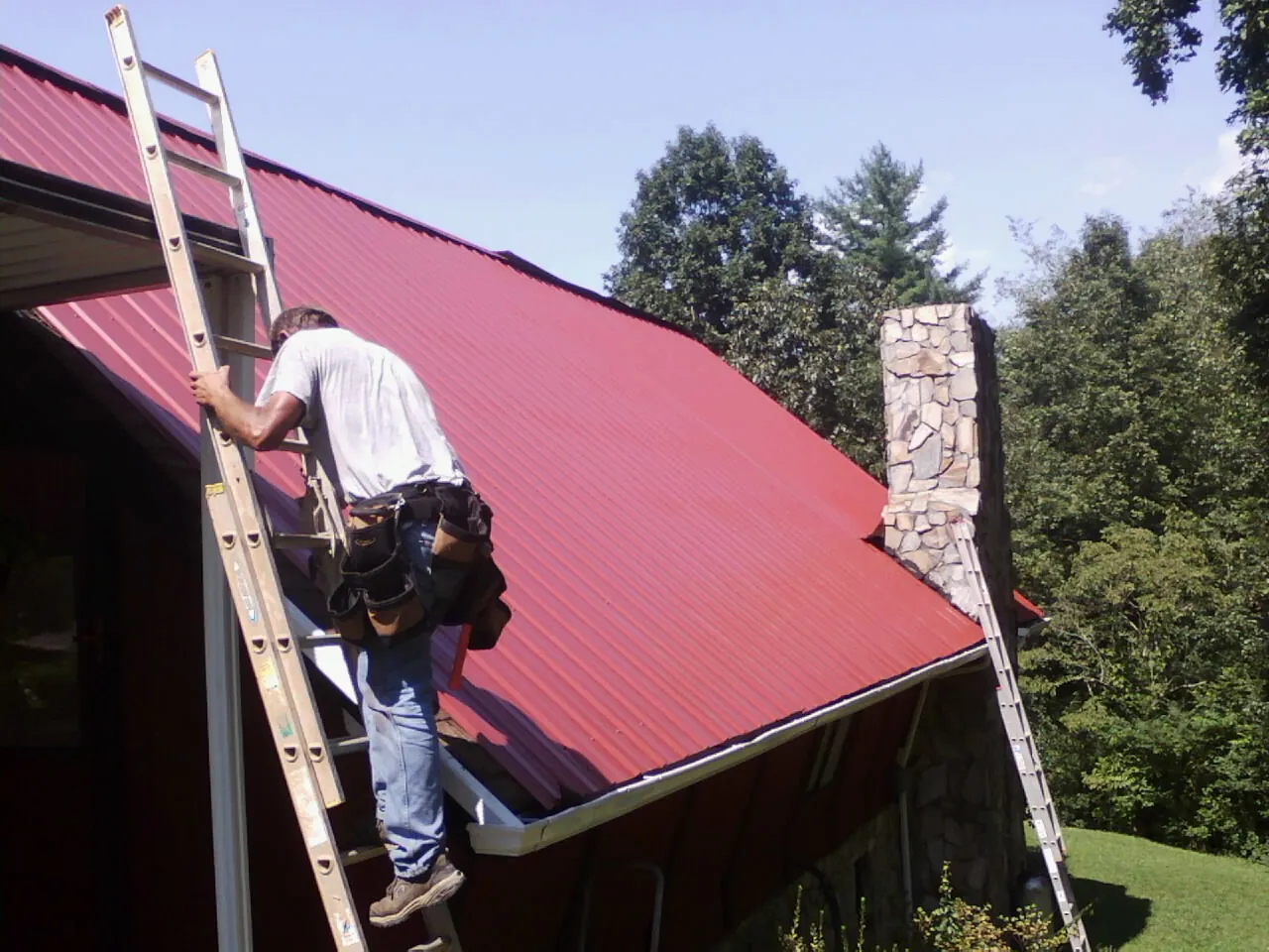 Roofer on a ladder beside a red metal roof and stone chimney — Soffit & Fascia Repair by Roofing Brothers in Westport